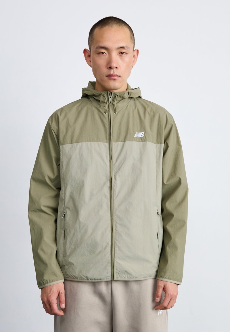 Veste à capuche vert clair avec une partie inférieure grise, fabriquée en matériau léger. Dotée d'une fermeture éclair à l'avant et de poches latérales. Logo New Balance affiché.