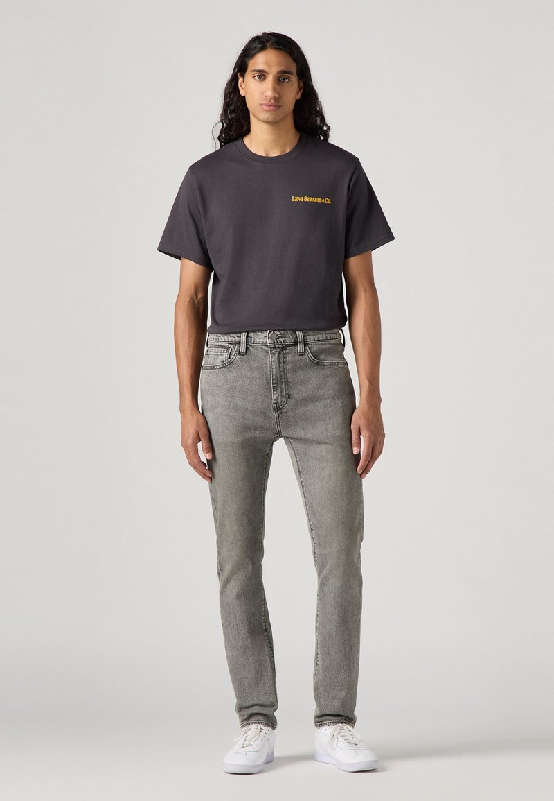 T-shirt grise avec logo jaune associée à un jean gris clair. Le modèle porte des baskets blanches. Design simple et décontracté avec une coupe ample.
