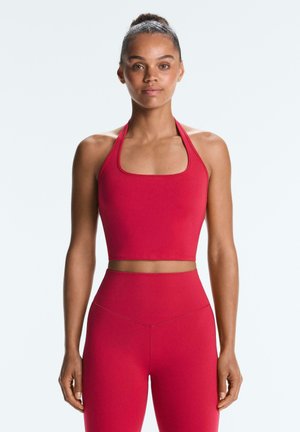 Žena s kosom u punđi, odjevena u crveni halter top i crvene tajice s visokim strukom, stoji ispred jednostavne svijetle pozadine.