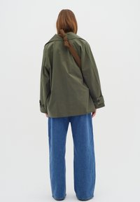 Giacca oversize verde oliva con colletto ripiegato, polsini abbottonati e una cintura marrone annodata. Indossata sopra jeans a gamba larga di un blu chiaro.