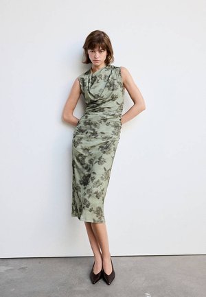 Femme en robe midi verte à fleurs sans manches et talons pointus noirs, debout contre un mur blanc avec les mains derrière le dos.