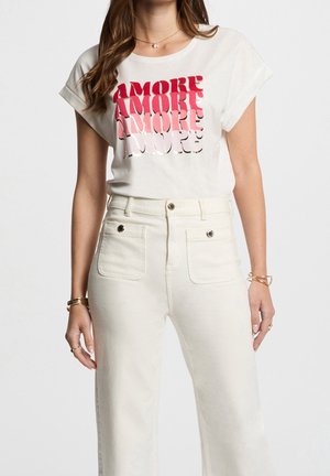 Frau trägt weißes T-Shirt mit hochgekrempelten Ärmeln und rotem Farbverlauf "AMORE"-Text sowie hoch taillierte weiße Hose mit vorderen Taschen mit Knöpfen.