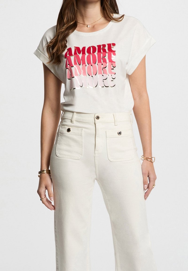 Femme portant un t-shirt blanc à manches retroussées avec un texte en dégradé rouge "AMORE" et un pantalon blanc taille haute avec poches avant à boutons.