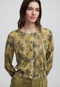 Blusa de manga larga abotonada en estampado de serpiente texurizado en amarillo y marrón. Presenta un escote redondo y botones oscuros en la parte delantera.
