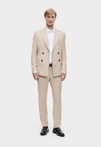 Suit beige doppiopetto con bottoni scuri, camicia bianca e scarpe nere. Il tessuto appare liscio, con una vestibilità adattata e pantaloni dritti.