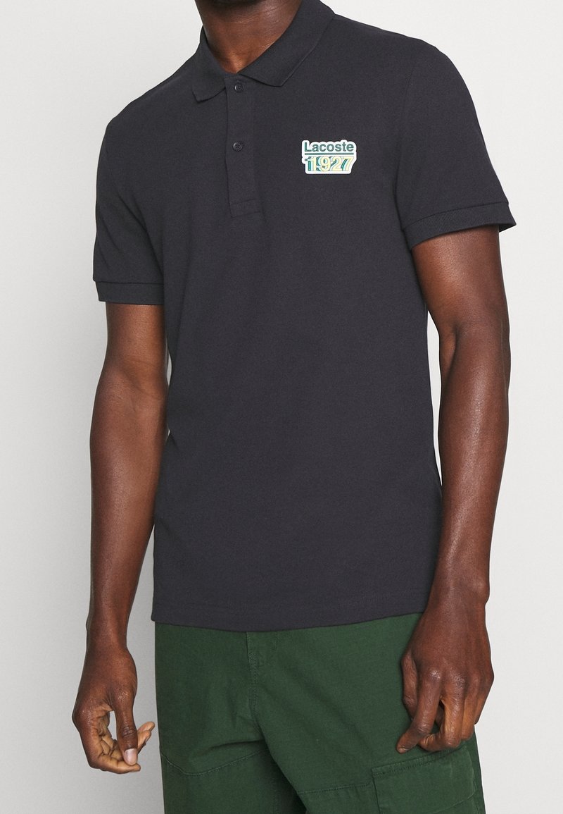 Homme portant un polo noir Lacoste avec le logo « Lacoste 1927 » sur la poitrine et un pantalon vert foncé, debout devant un fond uni.