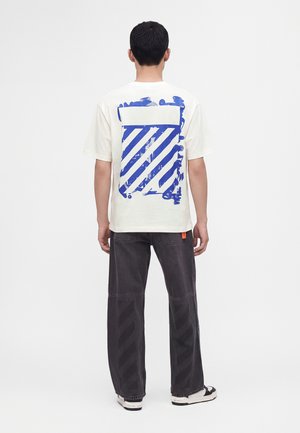 Mann trägt weißes T-Shirt mit großem blauen diagonalen Streifenmotiv auf dem Rücken, schwarze weit geschnittene Hose und weiße Sneakers, steht mit dem Rücken zum Betrachter.