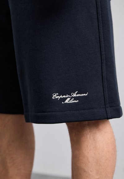 Tamsiai mėlyni sportiniai šortai, pagaminti iš minkštos medžiagos, turintys tekstūruotą paviršių ir baltą siuvinėtą „Emporio Armani Milano“ logotipą ant apatinės dalies.
