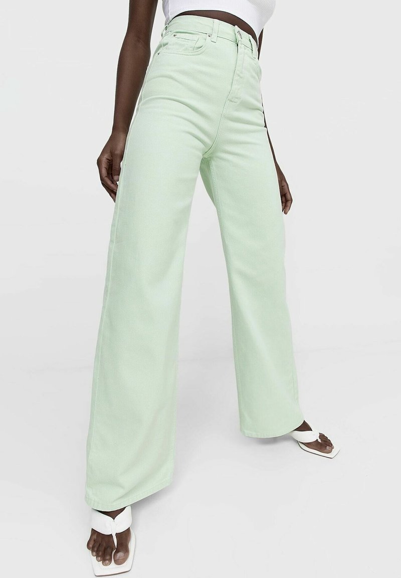 Personne portant un jean taille haute à jambes larges vert pastel, associé à des sandales blanches à bout carré et un top blanc court, debout sur un fond blanc.