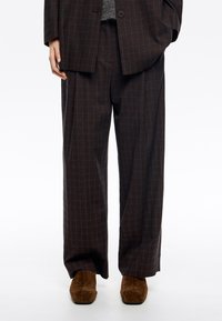 Pantaloni a quadri marroni realizzati in un tessuto testurizzato, con una vestibilità ampia e vita alta. Abbinati a scarpe in suede marrone.