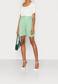 Maglietta grigia chiara con scollo a barca abbinata a pantaloni corti in lino verdi. La modella tiene una borsa verde scuro e indossa scarpe con tacco bianche.
