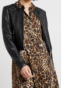 Žena v černé kožené bundě přes košilové šaty s leopardím potiskem, ruka opřená blízko pasu.