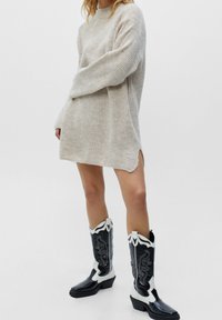 Robe pull en tricot beige avec une texture côtelée et des fentes latérales, associée à des bottes de cowboy noires et blanches montantes ornées de motifs détaillés.