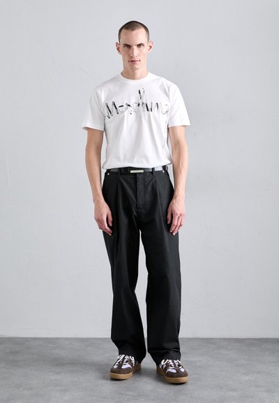 Tricou alb cu un design imprimat, pantaloni negri și adidași maro cu accente albe. Material texturat și croială lejeră într-un cadru neutru.