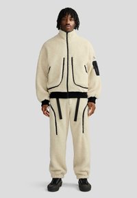 Giacca in pile color crema con tasche con zip, dettagli neri, polsini a costine e pantaloni abbinati con cinghie e dettagli con zip.