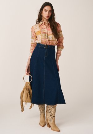 Frau trägt eine pinke Patchworkbluse, einen hoch taillierten dunkelblauen Denim-Midirock, beige Stiefeletten mit Blumenmuster und Cut-outs, hält eine braune Fransenhandtasche.