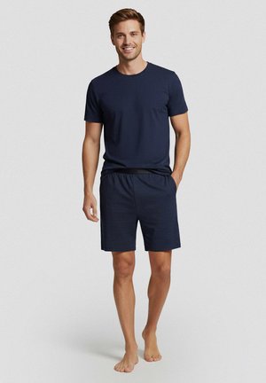 Lachende man zonder schoenen staand, met een donker marineblauw T-shirt met korte mouwen en bijpassende elastische taille korte broek tegen een effen lichte achtergrond.