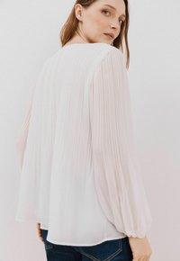 Lys pink, plisseret bluse med lange, samlede ærmer, tekstureret stof og rund halsudskæring, parret med mørke denim jeans.