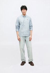 Ανοιχτό μπλε ριγωτό πουκάμισο με κουμπιά και λογότυπο στο στήθος, συνδυασμένο με χαλαρά ανοιχτόχρωμα τζιν και καφέ παπούτσια. Το μοντέλο στέκεται στο κέντρο.