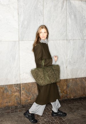 A woman walks against a marble wall, wearing an olive coat and gray shirt, carrying a textured green handbag, with black sneakers on her feet.