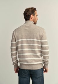 Man facing away wearing beige knit sweater with white horizontal stripes and blue jeans against neutral background.