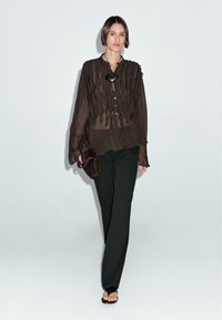 Donna che cammina con una blusa nera trasparente con dettagli in frange, pantaloni neri, collana con pendente nera, mentre porta una borsa nera e sandali neri scoperti.