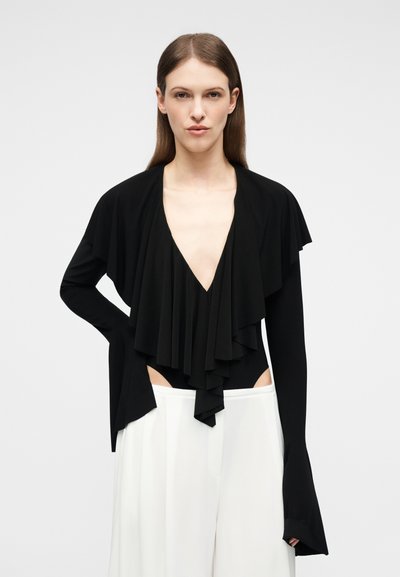 Γυναίκα με μακριά καστανά μαλλιά φοράει μαύρο τοπ με βαθύ V ντεκολτέ και στρώσεις μπροστά, με μακριά μανίκια, σε συνδυασμό με λευκό φαρδύ παντελόνι ψηλόμεσο.