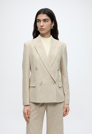 Femme aux longs cheveux foncés porte un blazer beige à double boutonnage et un pantalon assorti, accessoirisés de boucles d’oreilles dorées en forme d’anneaux, regardant vers la gauche.