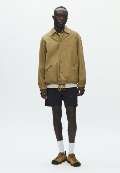 Homme noir portant une veste boutonnée couleur sable, un short noir, des chaussettes blanches et des chaussures de randonnée couleur sable, debout devant un fond blanc uni.