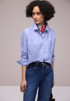 Mujer con cabello rizado que lleva una camisa azul clara abotonada, bufanda roja a rayas, vaqueros azul oscuro y sostiene un bolso negro.