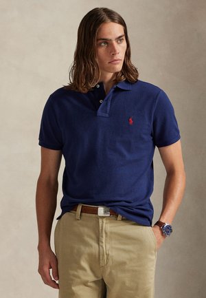 Ung mann med langt hår iført en mørkeblå poloshorte med rødt logo, beige bukser, brun belte og blå armbåndsur, stående med hånden i lommen.