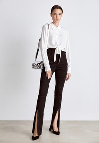 Camicia bianca a maniche lunghe con nodo sul davanti, pantaloni neri a vita alta con spacchi laterali e tacchi neri. Borsa tracolla argento che aggiunge contrasto.