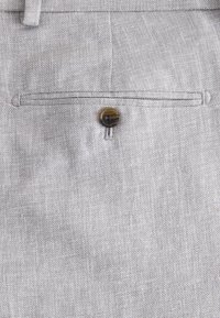 Gros plan d’un pantalon en tissu beige avec une poche arrière boutonnée, une couture nette et une passant de ceinture en haut.