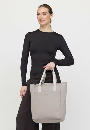 Frau trägt langärmeliges schwarzes Oberteil und Hose und hält eine große beige Tragetasche mit doppelten Griffen vor einfachem Hintergrund.