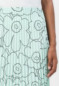 Mintgrønn plisserte skjørt med et svart blomstermønster. Glatt tekstur med elastisk midje. En hånd ser ut til å holde stoffet.