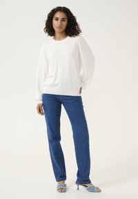 Witte gebreide trui met textuur, poffende mouwen en een ronde halslijn, gecombineerd met blauwe recht-toe-rechtdoor jeans en lichtblauwe haksandalen.