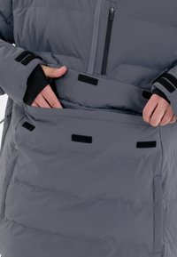 Grå isoleret jakke med en lynlåslomme og velcro-lukninger. Har en glat tekstur og et fyldigt design med vandrette syninger.