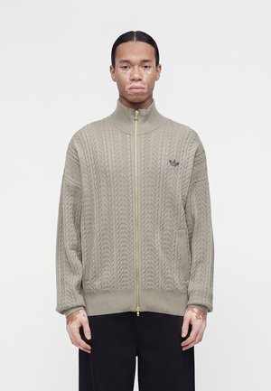 Persona con vitíligo vistiendo un jersey beige con textura y cremallera con logo de Adidas, de pie frente a un fondo blanco liso.