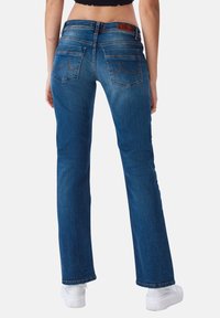Pantalones vaqueros acampanados de mezclilla azul con dos bolsillos traseros, ligera decoloración y costuras de contraste, combinados con zapatillas blancas.