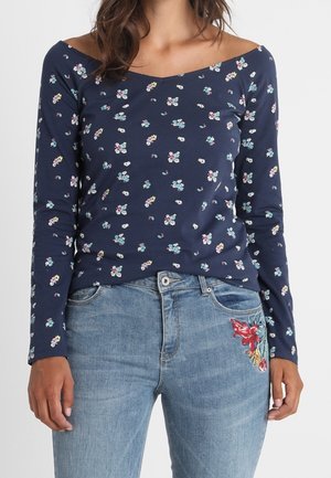 Žena oblečená v námořnicky modrém květovaném topu s dlouhým rukávem a odkrytými rameny a světle modrých džínách s červenou motýlí výšivkou na kapse.