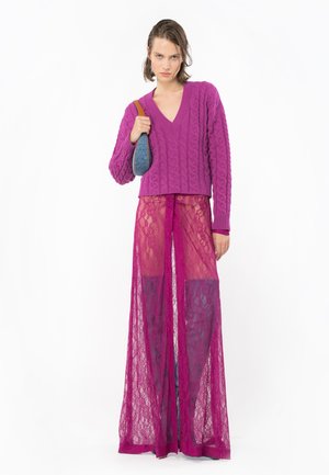 Suéter de punto morado brillante con escote en V y diseño de trenzas, combinado con pantalones de encaje rosa transparentes de piernas anchas.