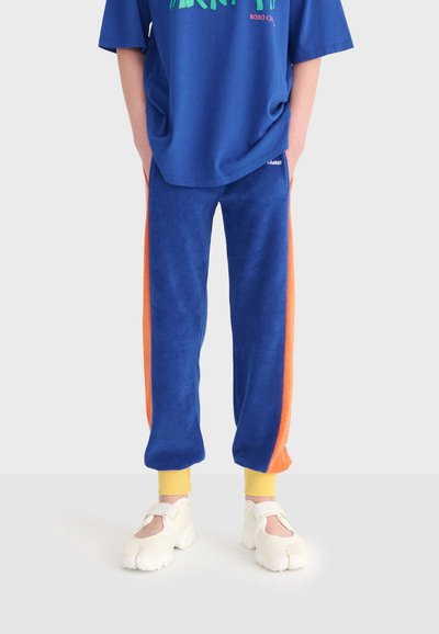 Persona che indossa pantaloni blu con strisce laterali arancioni, polsini gialli, una t-shirt grafica blu e scarpe bianche con punta divisa, in piedi contro uno sfondo chiaro.