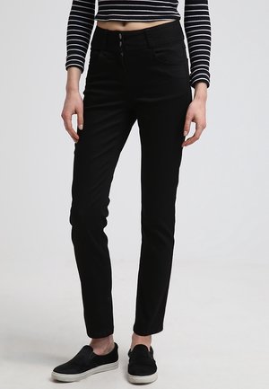 Kobieta ubrana w czarne dopasowane spodnie, czarne wsuwane buty oraz czarno-biały prążkowany krótki top z długim rękawem, stoi na jednolitym tle.