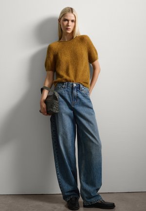 Suéter de punto mostaza de manga corta, jeans azules holgados y un bolso gris texturizado con asa, todo sobre un fondo neutro.