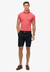 Polo shirt rossa con colletto, abbinata a pantaloni corti blu navy. Il modello indossa una cintura marrone e scarpe marroni con lacci. Design semplice e casual.