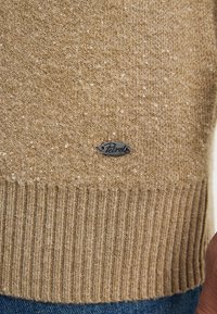 Gouden gebreide trui met een geribde zoom, subtiele sprankelende textuur en een klein metalen merklabel aan de onderzijde aan de linkerkant.