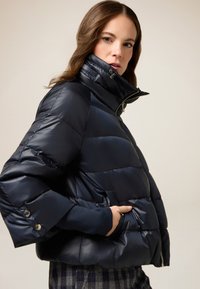 Mørkeblå pufferjakke med høj krave, lynlåslukning og sidelommer. Har vandret quiltet mønster og ribbede manchetter.