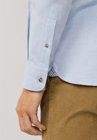 Camisa de botón azul claro de manga larga. Los puños tienen botones marrones y un patrón a cuadros debajo del dobladillo. Combinada con pantalones de pana.