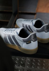 Witte sneakers met grijze strepen, gestructureerd synthetisch materiaal, beige rubberen zool en een prominent logo-label op de hiel.