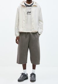 Person iklädd en ljus beige huvtröja, en vit grafisk t-shirt med mesh, grå vida cropped byxor, vita strumpor och mörkgrå sneakers på vit bakgrund.